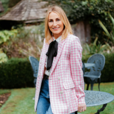 Mel Cheeseman in a pink checkered jacket standing outdoors with greenery in the background